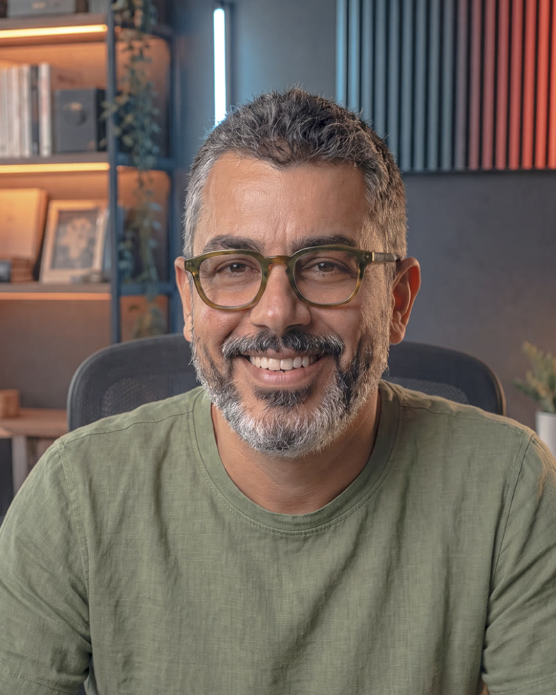 Bruno Hilel sorrindo, de óculos e camiseta verde, no estúdio dele